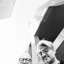Selfie von Roland Puck mit einem Freund vor moderner Architektur.
