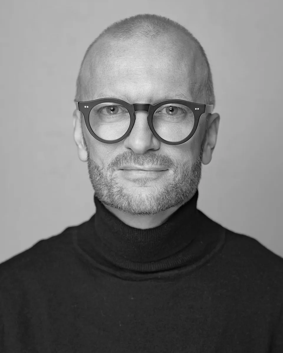 Roland Puck in Schwarz-Weiß, mit runder dunkler Brille und schwarzem Rollkragenpullover.
